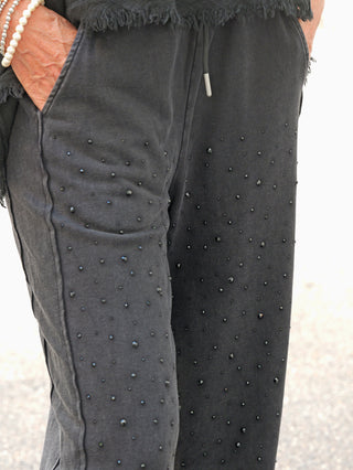 Die OFF#DLY Joggpant Daria black washed zeigt glitzernde Nieten auf schwarzen Joggerhosen, mit elastischem Bund und Eingrifftaschen. Eine Hand trägt ein Perlarmband.