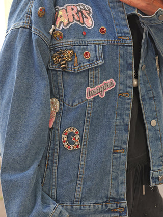 Nahaufnahme der OFF#DLY Denim Jacket Viola midblue mit Patches: „ARTS“, „Imagine“, ein Bienenbrosche, ein Strass-„G“ und Perlenherz, getragen über dunklem Hoodie.