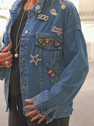 OFF#DLY Denim Jacket Viola midblue zeigt eine oversized Jeansjacke mit dekorativen Ansteckern und Patches wie einem glitzernden Stern, einer Imagine-Stickerei und einer 1970er-Jahreszahl, getragen über einem schwarzen Top.