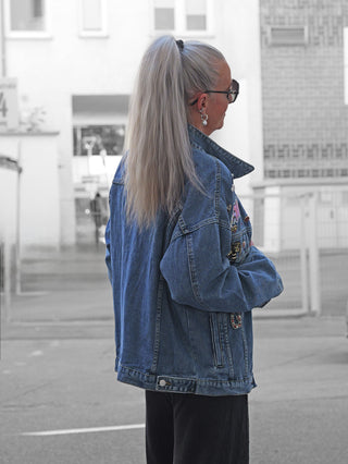 Person von hinten in OFF#DLY Denim Jacket Viola midblue mit Patches. Die Jeansjacke hat weite Ärmel und einen lässigen Schnitt, getragen in urbaner Umgebung.