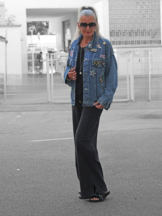 Frau trägt OFF#DLY Denim Jacket Viola midblue, verziert mit Patches und Ansteckern, über schwarzem Outfit. Accessoires: Sonnenbrille, Ohrringe, Kette. Hintergrund: Tor und Gebäudefassaden.