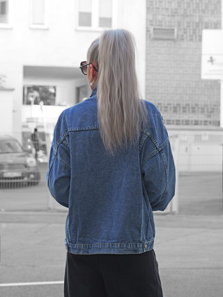 Person mit dem Rücken zur Kamera trägt die OFF#DLY Denim Jacket Viola midblue, eine locker geschnittene Jeansjacke mit auffälligen Patches und Stickern, vor urbaner Kulisse.