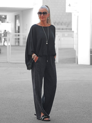 Frau trägt OFF#DLY Joggpant Daria black washed, eine lockere, schwarze Hose mit funkelnden Glitzernieten, kombiniert mit flachen Sandalen und einer silbernen Halskette, präsentiert einen entspannten, modischen Look.