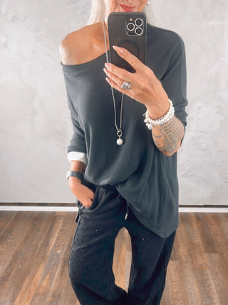 Person in schwarzem, schulterfreiem OFF#DLY Shirt Elli anthrazit, kombiniert mit weißen Ärmeln und schwarzer Hose mit Nieten. Silberne Kette mit Perle und weiße Armbänder ergänzen den Look.