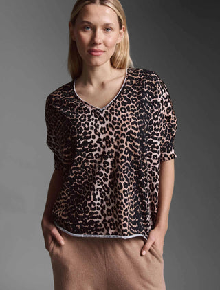 Frau trägt das Cotton Candy Tee Helay multicolor, ein locker sitzendes, kurzärmeliges Leoparden-T-Shirt mit V-Ausschnitt und Saumversatz, kombiniert mit hellbraunen Hosen. Hände in Hosentaschen.