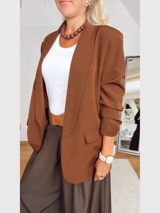 Frau trägt den OFF#DLY Blazer Chiara zimt mit entspannter Passform, hochgeschobenen Ärmeln. Kombiniert mit weißem T-Shirt, brauner Halskette und braunem Gürtel für einen eleganten, lässigen Look.