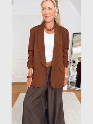 Frau im OFF#DLY Blazer Chiara zimt, rustbraun, mit kastigem Schnitt. Getragen über weißem Oberteil, kombiniert mit breiten, dunklen Hosen. Accessoires: Holz-Perlenkette, Armband und brauner Gürtel.