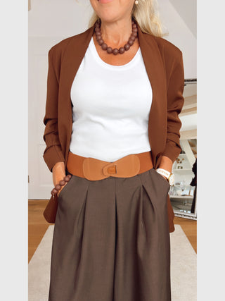 Person trägt den OFF#DLY Blazer Chiara zimt, lässig über die Schultern drapiert, kombiniert mit einem weißen Top und braunen, hochtaillierten Hosen, betont durch einen breiten, elastischen Gürtel.