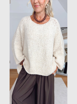 Person trägt BSB Pullover Ivoire, einen locker gestrickten, cremefarbenen Pullover mit überschnittenen Schultern und weiten Ärmeln. Dazu trägt sie breite, dunkle Hosen und klobige braune Perlenaccessoires.