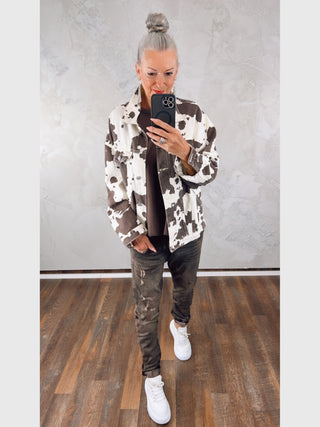 Person in Jeansjacke „Noemi“ beige/braun mit Kuhflecken-Print, dunklem Top, dunklen, leicht zerstörten Jeans und weißen, klobigen Sneakers steht auf Holzfußboden und macht ein Spiegel-Selfie.