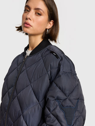 Junge Frau trägt eine ALIX Quilted Bomber navy mit Rautensteppung und Stierpatch auf dem Ärmel, silberne Creolen und ein helles Shirt darunter sind sichtbar.