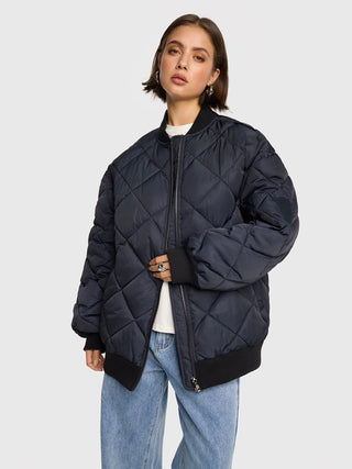 Junge Frau trägt die ALIX Quilted Bomber navy, eine übergroße, gesteppte Bomberjacke mit Reißverschluss, Rippenbündchen und Stickerei am Ärmel, kombiniert mit einem weißen Top und hellen Jeans.