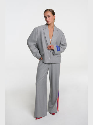 Frau trägt den ALIX Coated Blazer soft grey melange, einen übergroßen Blazer mit Fischgrätenmuster und The Label-Sticker am Ärmel, kombiniert mit passenden weiten Hosen mit Rennstreifen.