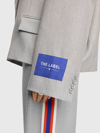 Nahaufnahme des ALIX Coated Blazer in soft grey melange, mit „THE LABEL”-Patch am Ärmel und rot-weiß-blauem Streifen an der Hose. Trägerhand mit schlichtem Silberring sichtbar.