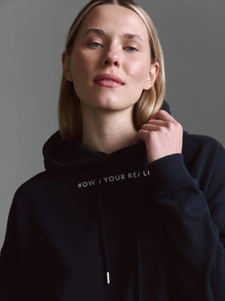 Junge Frau trägt das Cotton Candy Sweatshirt Malve black mit Kapuze, zieht am Kragen, sichtbarer Slogan-Teil OWN YOUR REAL. Lässige Passform, ideal für vielseitige Kombinationen.