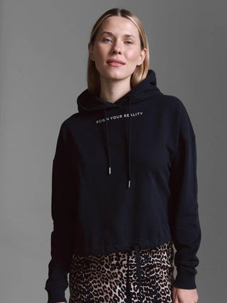 Person trägt das Cotton Candy Sweatshirt Malve black mit Kapuze und Tunnelzug, bedruckt mit OWN YOUR REALITY auf der Brust, kombiniert mit leopardengemusterten Hosen.