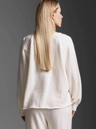 Rückansicht einer Frau im Cotton Candy Sweatshirt Eloa offwhite mit V-Ausschnitt, Ballonärmeln und Blusenmanschette, kombiniert mit passenden, locker sitzenden Hosen, vor neutralem Hintergrund.