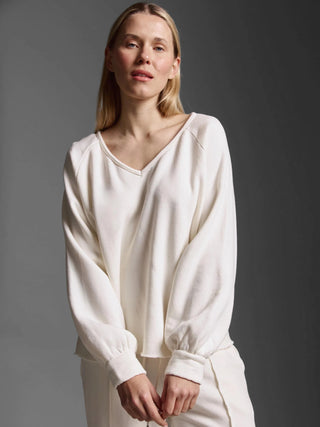 Eine Frau trägt das Cotton Candy Sweatshirt Eloa offwhite, ein locker sitzendes, offwhite Langarmshirt mit V-Ausschnitt und Ballonärmeln, kombiniert mit passenden Loungehosen, vor neutralem Hintergrund.