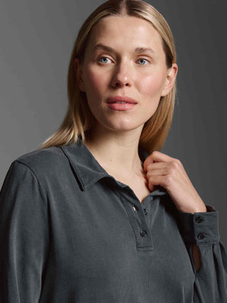 Nahaufnahme einer jungen Frau mit blondem Haar in einem dunkelgrauen Hemd, die den Kragen berührt. Produktfokus: Cotton Candy Dress Magnolia black, ein gewaschenes Kleid mit Polokragen und Seitenschlitzen.