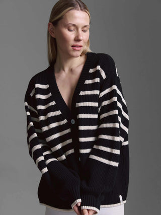 Person trägt Cotton Candy Cardigan Kristi, einen schwarz-cremeweiß gestreiften Oversized-Strickcardigan mit V-Ausschnitt, Knöpfen und leicht übergroßen Ärmeln mit gerippten Bündchen.