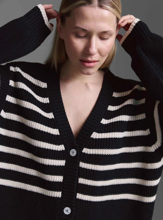 Frau trägt den Cotton Candy Cardigan Kristi, einen groben, schwarz gestreiften Oversized-Strickcardigan mit tiefem V-Ausschnitt und zwei großen Knöpfen, während sie ihre Haare richtet.