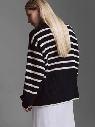 Modell von hinten in Cotton Candy Cardigan Kristi, gestreifter oversized Strickcardigan mit schwarzen und cremefarbenen Streifen oben, schwarzer unterer Teil, über einem hellen Rock oder Kleid, grauer Hintergrund.