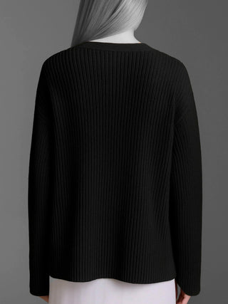 Eine Person trägt den Cotton Candy Cardigan Kristi black mit lockerem, oversized Schnitt, vertikalem Rippmuster und besonderen Ärmelabschlüssen. Ein weißer Rocksaum ist unter dem Pullover erkennbar.