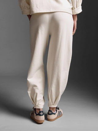Person von hinten in Cotton Candy Pant Giana offwhite, weite Passform, mit einzigartigem Beinsaum und seitlichen Taschen, kombiniert mit Adidas Sneakers, sichtbar vor neutralem Hintergrund.