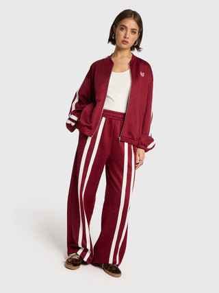 Junge Frau trägt ALIX Training Sweat Pant bordeaux mit weißen Streifen, kombiniert mit passender Jacke, weißem Tanktop und leopard-print Slip-On-Schuhen. Weite Hose aus Baumwoll-Polyester-Mix mit elastischem Bund.