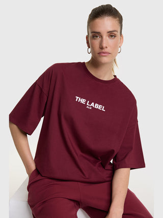 Eine junge Frau trägt das ALIX Basic The Label Tee bordeaux mit „THE LABEL“-Aufdruck in Weiß, entspannter Passform und Rundhalsausschnitt, kombiniert mit passenden burgunderfarbenen Hosen.