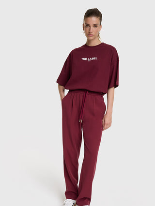Frau trägt das ALIX Basic The Label Tee bordeaux, ein lockeres, oversized T-Shirt mit „THE LABEL ALIX“ Aufdruck, kombiniert mit passenden Jogginghosen. 100% Baumwolle, entspannte Passform, Rippbündchen.