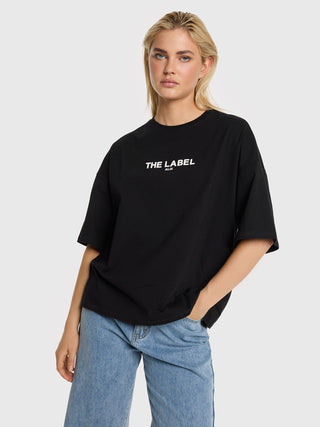 Junge Frau trägt das ALIX Basic The Label Tee black mit weitem Schnitt, „THE LABEL“-Aufdruck, fallenden Schultern, ellenbogenlangen Ärmeln, kombiniert mit gerade geschnittenen, hellblauen Jeans.