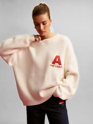 Junge Frau im ALIX Basic Jumper cream: Oversized, gerippter Strickpullover mit rotem A und THE LABEL-Schriftzug auf der Brust. Kombiniert mit dunkel gewaschener Jeans, rotem Logo-Patch.