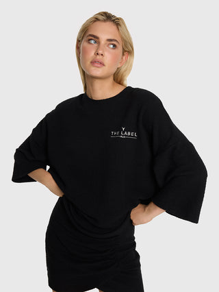 Junge Frau in ALIX Boucle Tee black: übergroßes schwarzes Bouclé-Jersey-Shirt mit „THE LABEL”-Stickerei, dreiviertel Ärmel, kombiniert mit eng anliegendem schwarzem Rock, auf hellem Hintergrund.
