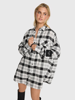 Eine junge Frau trägt das ALIX Check Blouse in Schwarz/Weiß, ein oversized, langärmeliges, karriertes Hemd mit Brusttasche und Label am Ärmel, kombiniert mit kniehohen schwarzen Stiefeln.