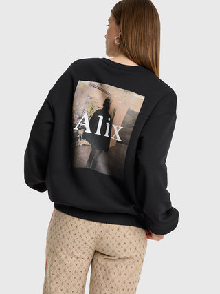 Eine Frau trägt den ALIX Photoprint Sweater black mit urbanem Fotodruck auf der Rückseite, kombiniert mit beigen Hosen mit Monogramm-Muster und roten Streifen.