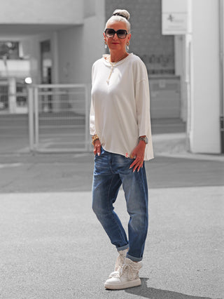 Person trägt das OFF#DLY Shirt Ciao Bella white, ein lässiges, locker sitzendes weißes Oberteil, kombiniert mit blauen Jeans und weißen Sneakers, ergänzt durch goldfarbene Schmuckakzente und schwarze Sonnenbrille.