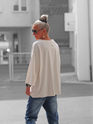 Ältere Frau in urbanem Umfeld trägt das OFF#DLY Shirt Ciao Bella white, ein oversized weißes Langarmshirt, kombiniert mit blauen Jeans, Sonnenbrille und Schmuck, verkörpert lässigen Street-Style.