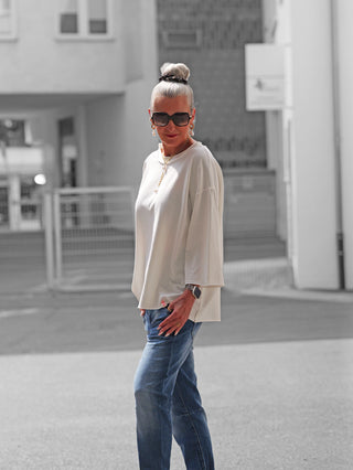 Frau in urbanem Umfeld mit OFF#DLY Shirt Ciao Bella white, lockerem Schnitt und dreiviertel Ärmeln, kombiniert mit blauer Jeans und Accessoires, darunter Sonnenbrille und Goldkette.