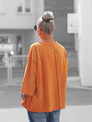 Ältere Frau von hinten in einem Oversize „Ciao Bella“ Shirt in Burnt-Orange, mit freiliegenden Nähten und lockeren Ärmeln, kombiniert mit blauen Jeans und modischem Accessoire.