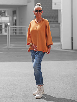 Ältere Frau trägt das OFF#DLY Shirt Ciao Bella orange, ein oversized Oberteil mit verkürzten Ärmeln, kombiniert mit blauen Jeans und weißen Sneakers, verleiht ihrem Look urbanen Chic.