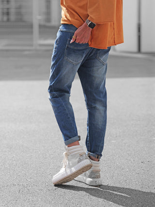 Person trägt OFF#DLY Denim Parma lightblue: schlank geschnittene, hochgekrempelte blaue Jeans im klassischen Five-Pocket-Stil, kombiniert mit weißen High-Top-Sneakers mit glitzernden Absätzen.