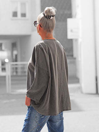 Person von hinten mit silbergrauem Dutt, trägt eine OFF#DLY Schlupfbluse Fiorella in Schwarz gewaschen, locker geschnitten, kombiniert mit blauen, farbbespritzten Jeans in urbaner Umgebung.