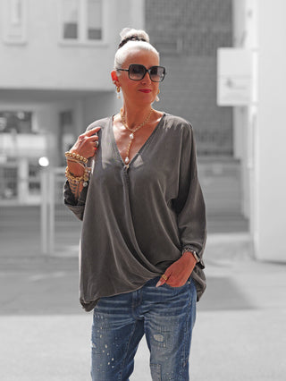Ältere Frau trägt die OFF#DLY Schlupfbluse Fiorella in Black Washed, locker sitzend mit V-Ausschnitt, kombiniert mit lässiger Jeans. Goldschmuck akzentuiert den eleganten Look in urbaner Umgebung.