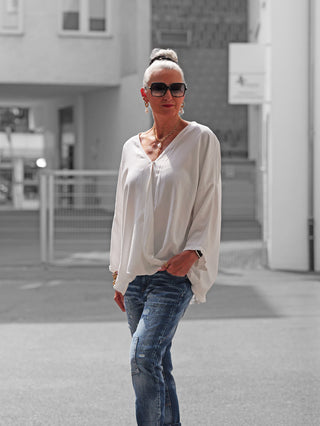 Frau in urbanem Hof trägt die OFF#DLY Schlupfbluse Fiorella in Weiß, kombiniert mit blauer Jeans. Accessoires: Sonnenbrille, Ohrringe, Goldschmuck. Bluse mit lockerem Schnitt, ideal für lässigen Stil.