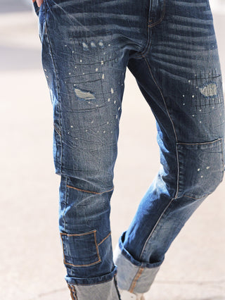 OFF#DLY Denim Livorno midblue: Beine in leicht oversized Jeans mit Distressed-Details, Patchwork-Panels und hochgekrempelten Bündchen, die das Innenmaterial zeigen, auf hellem Pflaster stehend.