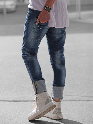 OFF#DLY Denim Livorno midblue: Slim-fit Jeans mit subtilen Paint-Splash-Details und schrägen Nähten, getragen mit hochgekrempelten Beinen. Kombiniert mit weißen High-Top-Sneakern und einem lässigen, übergroßen weißen Hemdsaum.