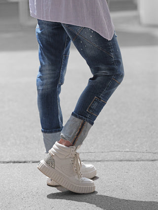 Nahaufnahme von Beinen in der OFF#DLY Denim Livorno midblue Jeans mit Patchwork-Details und Used-Effekten, kombiniert mit weißen, klobigen High-Top-Sneakern mit silbernen Akzenten.