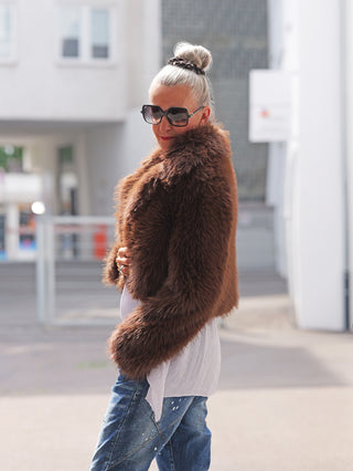Frau trägt auf der Straße die OFF#DLY Felljacke Luisa brown, eine kurz geschnittene, shaggy Kunstpelzjacke über weißer Bluse und blauen Jeans, ergänzt durch schwarze Sonnenbrille und Ringe.