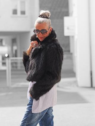 Die stilvolle, silberhaarige Frau trägt die OFF#DLY Felljacke Luisa black, einen cropped Kunstpelz, kombiniert mit lässigen Jeans und goldenen Schmuck, perfekt für trendbewusste Herbst- und Winterlooks.